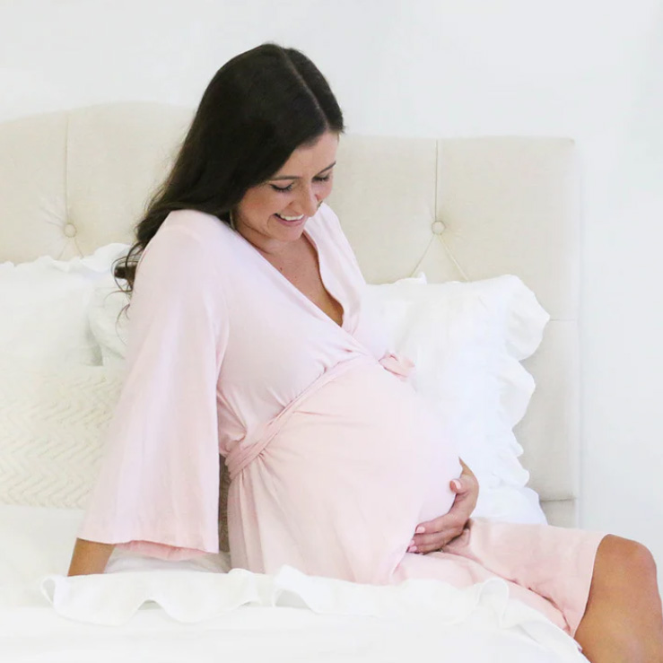 Expecting mother sitting on a bed wearing a pale pink bamboo knit maternity robe, holding her baby bump and smiling.